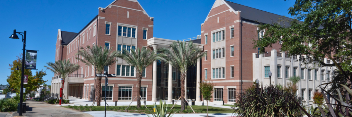 Florida State University’s Herbert Wertheim College of Business, a large brick academic building with tall windows, palm trees, and a central columned entrance, shown under a bright blue sky.